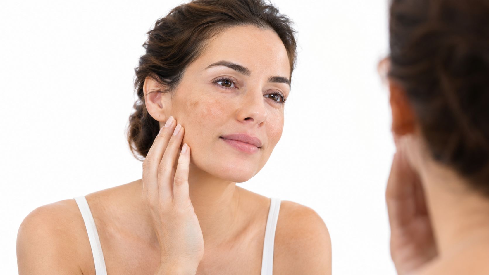 A woman examining hyperpigmentation on her cheek while looking in the mirror to check her skin condition.