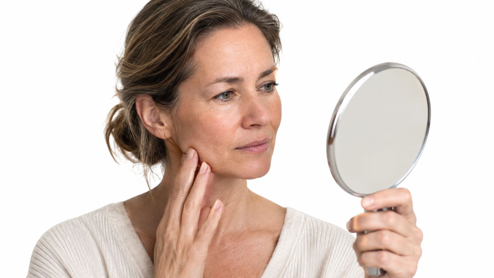 A middle-aged woman examining her face in a handheld mirror to check for signs of aging.